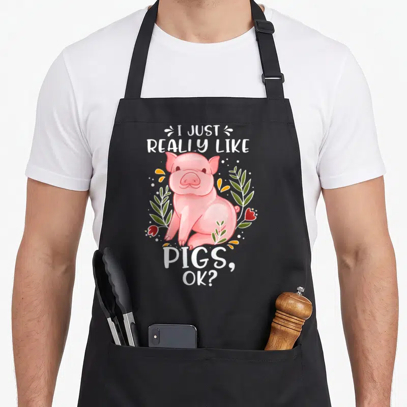 Retro pig lover apron - Pink cartoon pig surrounded by green leaves and red flowers with white text — shown on a black cotton canvas apron against a white background