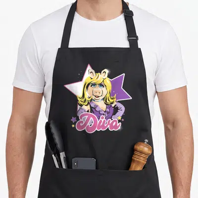 Diva little miss apron - Cartoon pig character in purple dress in front of a large star with pink Diva text — shown on a black cotton canvas apron against a white background