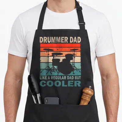 Drummer father day apron - Grey drum kit silhouette on red orange yellow teal grey striped background with retro text — shown on a black cotton canvas apron against a white background