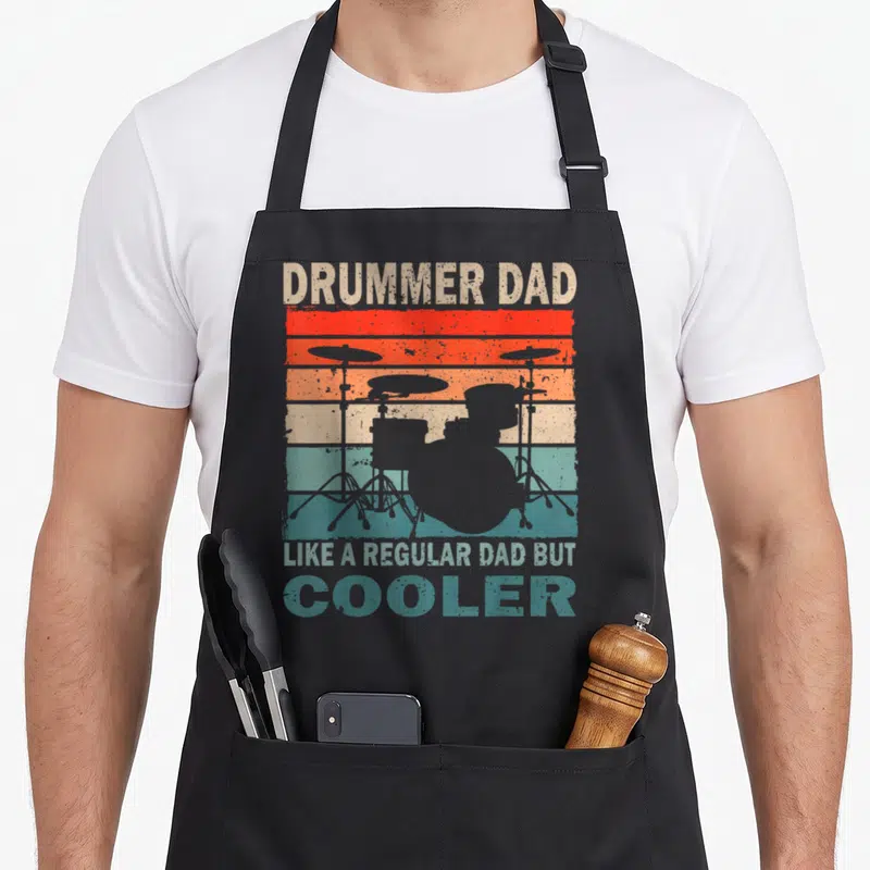 Drummer father day apron - Grey drum kit silhouette on red orange yellow teal grey striped background with retro text — shown on a black cotton canvas apron against a white background