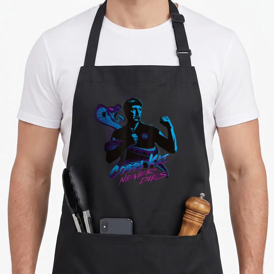 Kai mercy apron - Retro graphic of man and coiled cobra in blue purple pink with text Cobra Kai Never Dies — shown on a black cotton canvas apron against a white background