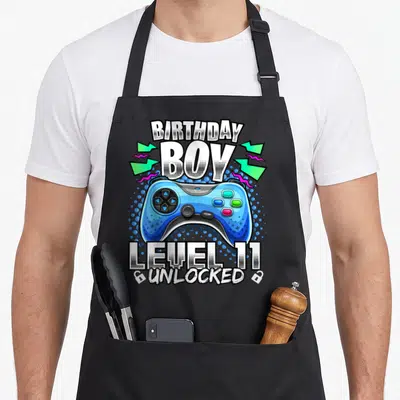 11th birthday video game apron - Blue game controller with BIRTHDAY BOY LEVEL 11 UNLOCKED text and lightning bolts — shown on a black cotton canvas apron against a white background