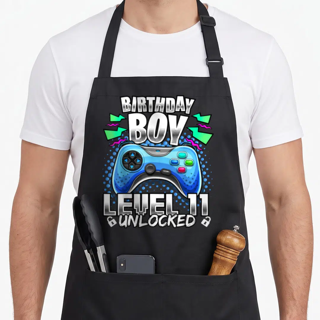 11th birthday video game apron - Blue game controller with BIRTHDAY BOY LEVEL 11 UNLOCKED text and lightning bolts — shown on a black cotton canvas apron against a white background