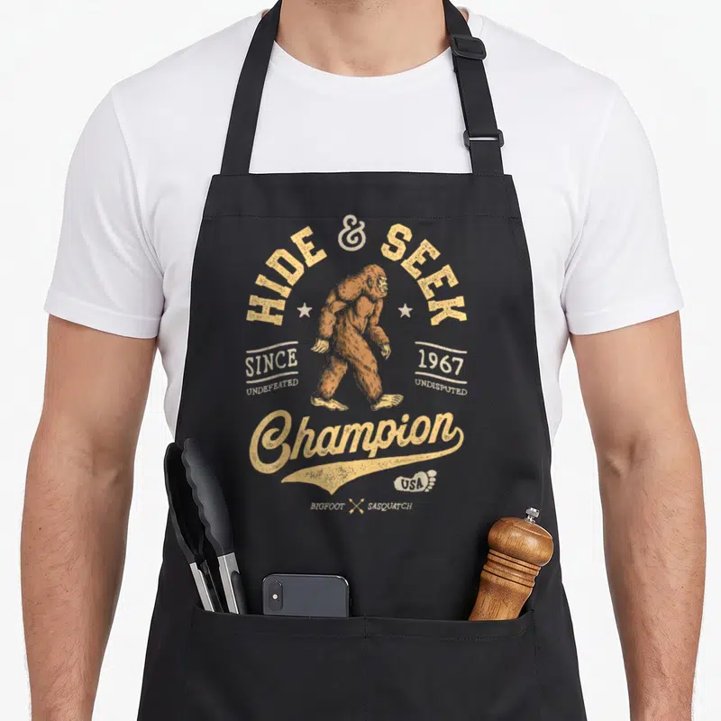 Hide seek champion bigfoot apron - Retro tan Bigfoot illustration with arching HIDE & SEEK text and 1967 dates — shown on a black cotton canvas apron against a white background