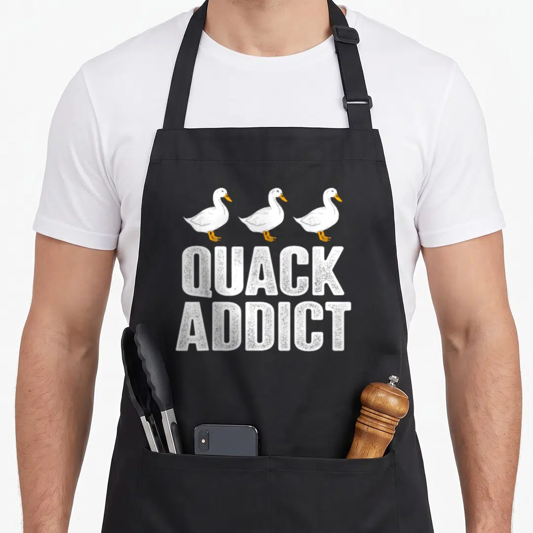 Quack duck apron - Three white ducks facing right above QUACK ADDICT stacked text — shown on a black cotton canvas apron against a white background