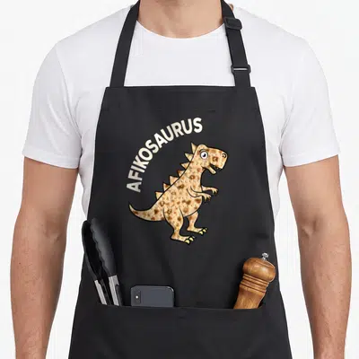 Dinosaur passover apron - AFIKOSAURUS text arching over a tan dinosaur shaped like matzah bread — shown on a black cotton canvas apron against a white background