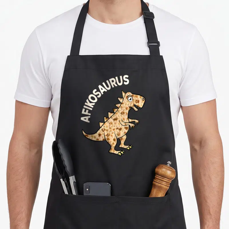 Dinosaur passover apron - AFIKOSAURUS text arching over a tan dinosaur shaped like matzah bread — shown on a black cotton canvas apron against a white background