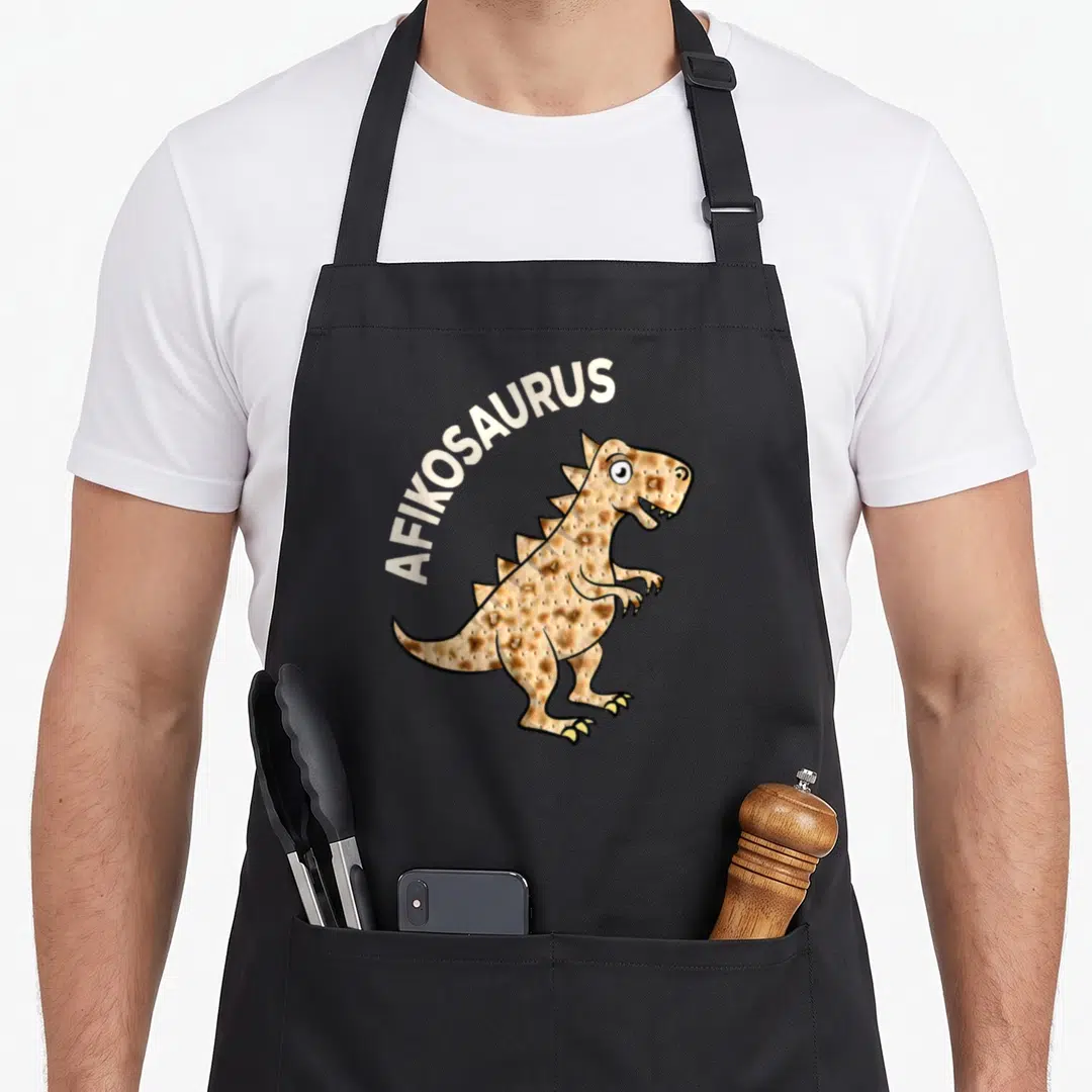 Dinosaur passover apron - AFIKOSAURUS text arching over a tan dinosaur shaped like matzah bread — shown on a black cotton canvas apron against a white background