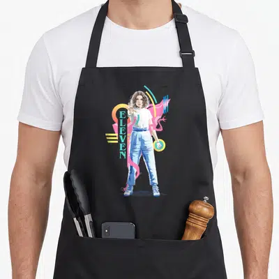 Eleven 80s apron - Retro character illustration with vertical ELEVEN text and pink yellow blue teal geometric shapes — shown on a black cotton canvas apron against a white background