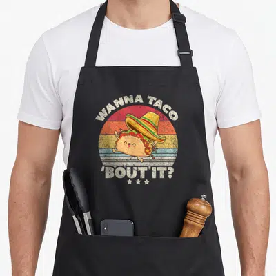 Funny bout taco apron - Retro taco wearing sombrero with WANNA TACO 'BOUT IT? text and striped sun background — shown on a black cotton canvas apron against a white background