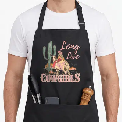 Live western apron - Long Live COWGIRLS text with cartoon cowgirl riding spotted horse near green cactus — shown on a black cotton canvas apron against a white background