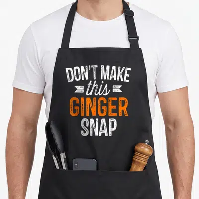 Redhead snap aprons - White and orange stacked text reading DON'T MAKE this GINGER SNAP with decorative banners — shown on a black cotton canvas apron against a white background