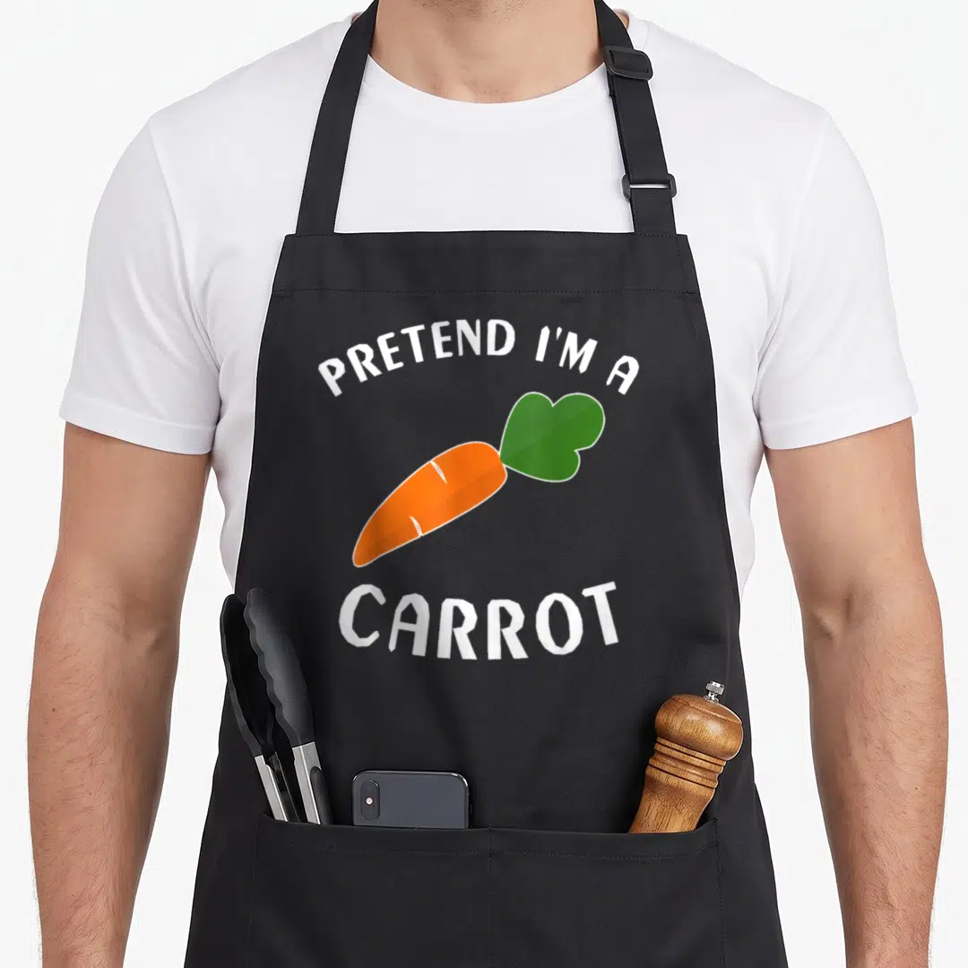 Carrot easy halloween apron - Cartoon orange carrot with green leaves and white text PRETEND I'M A CARROT — shown on a black cotton canvas apron against a white background