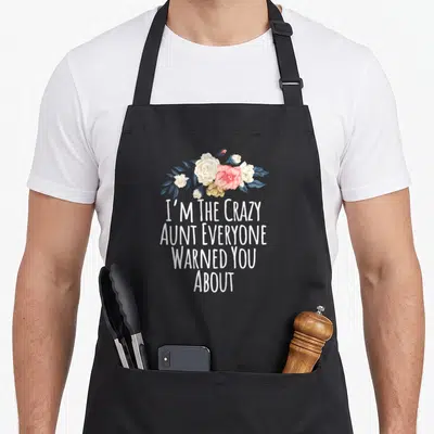 Funny aunt novelty apron - Floral bouquet above white text reading I'M THE CRAZY AUNT EVERYONE WARNED YOU ABOUT — shown on a black cotton canvas apron against a white background