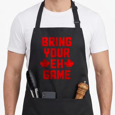 Bring pride apron - Red stacked text BRING YOUR EH GAME flanked by two maple leaves — shown on a black cotton canvas apron against a white background