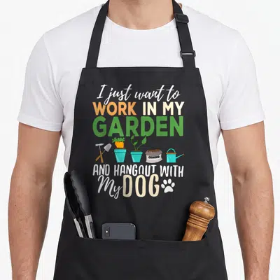 Gardening funny dog apron - I just want to work in my garden and hangout with my dog text with cartoon trowel, carrots — shown on a black cotton canvas apron against a white background