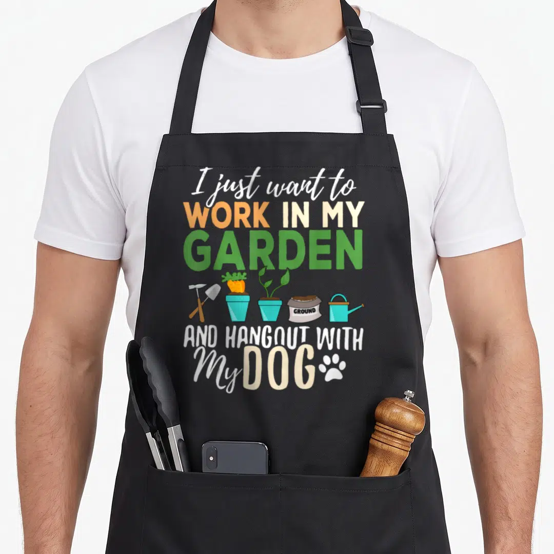 Gardening funny dog apron - I just want to work in my garden and hangout with my dog text with cartoon trowel, carrots — shown on a black cotton canvas apron against a white background