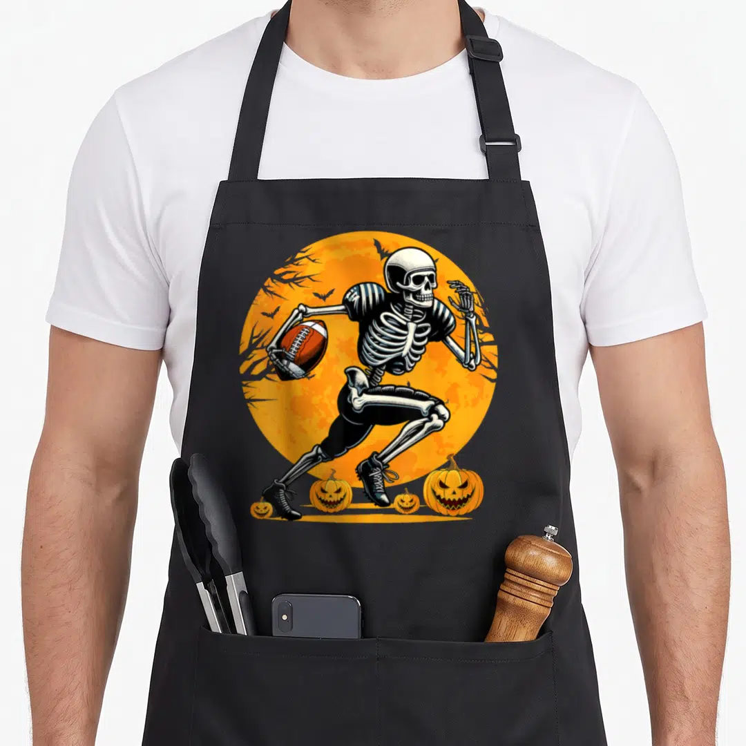 Football skeleton halloween apron - Cartoon skeleton running with football in front of orange moon and pumpkins — shown on a black cotton canvas apron against a white background