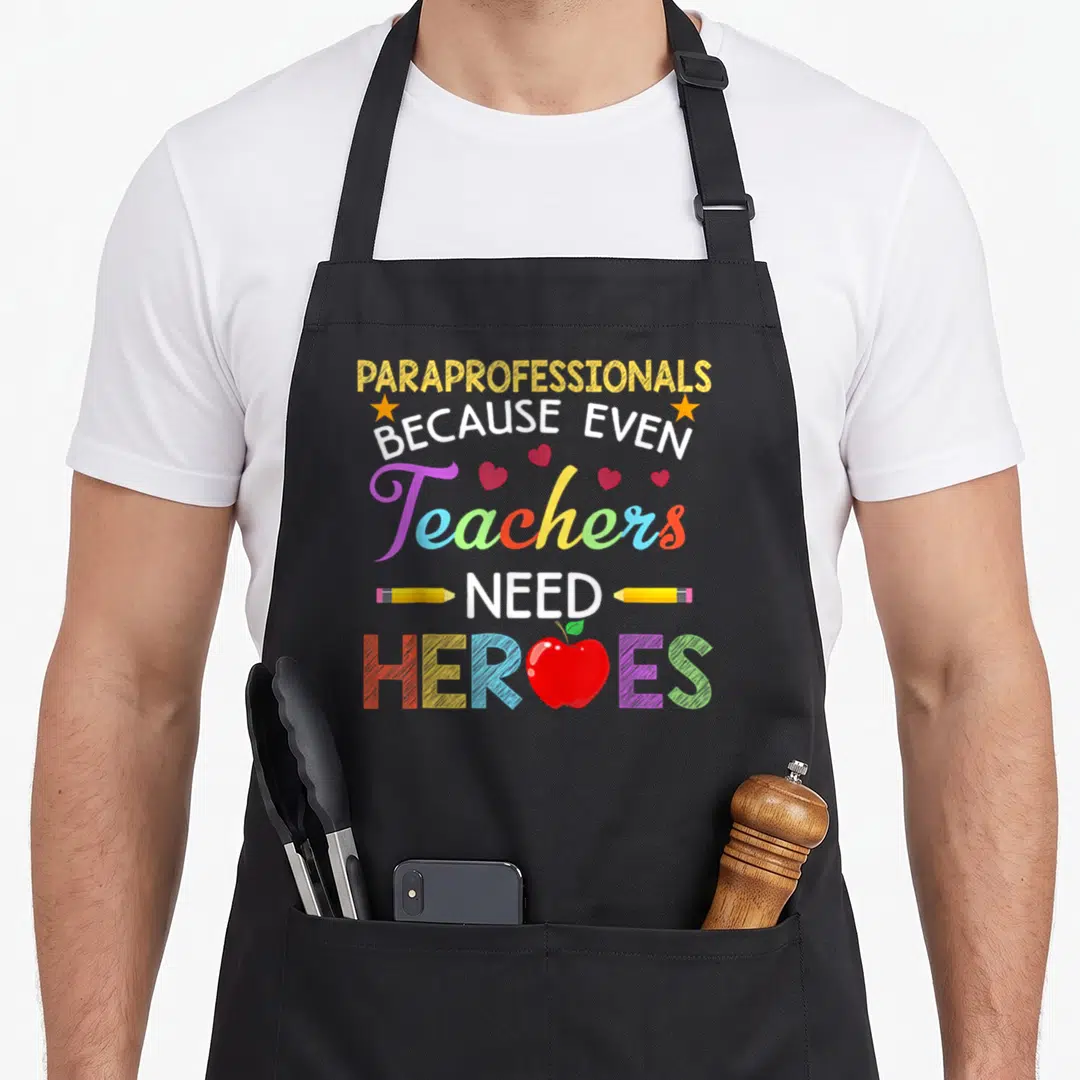 Paraprofessional teacher apron - Stacked colorful text with apple replacing O in HEROES and pencils flanking NEED — shown on a black cotton canvas apron against a white background