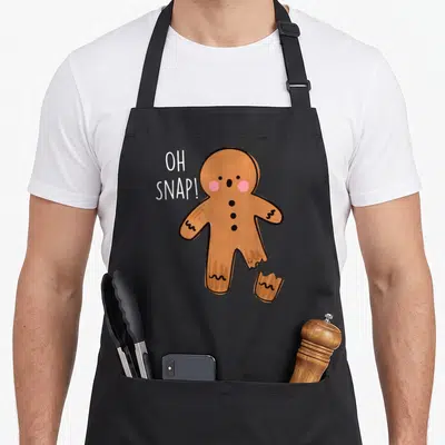 Bone gingerbread apron - Cartoon gingerbread man with broken leg and OH SNAP text in brown white pink — shown on a black cotton canvas apron against a white background