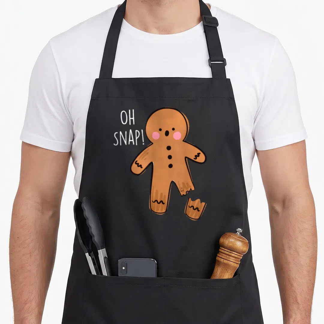 Bone gingerbread apron - Cartoon gingerbread man with broken leg and OH SNAP text in brown white pink — shown on a black cotton canvas apron against a white background