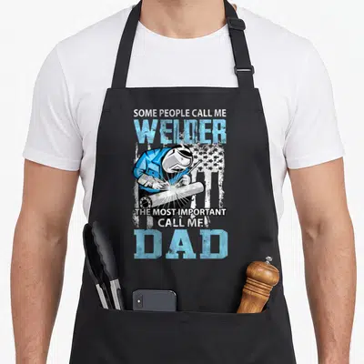 Welder daddy father day apron - Cartoon welder on distressed flag with blue and white text celebrating dad — shown on a black cotton canvas apron against a white background