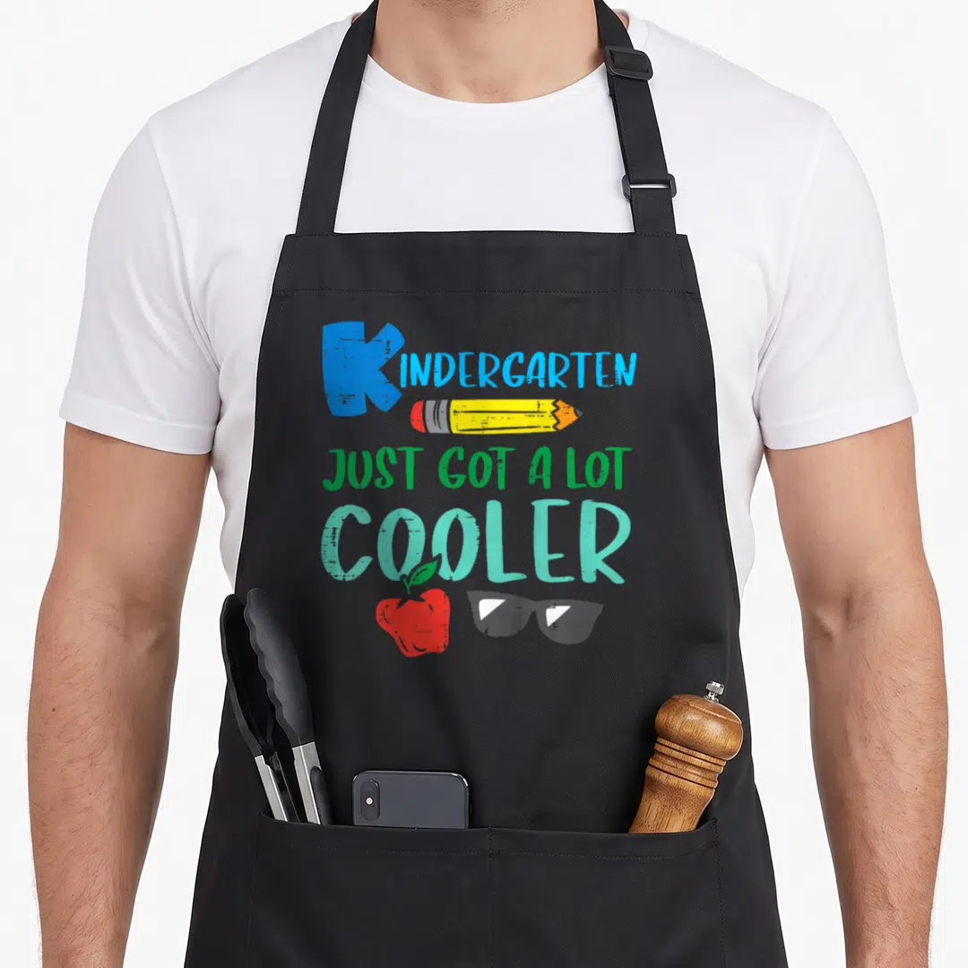 Cooler kindergarten apron - Stacked text reading KINDERGARTEN JUST GOT A LOT COOLER with pencil, apple, and sunglasses — shown on a black cotton canvas apron against a white background