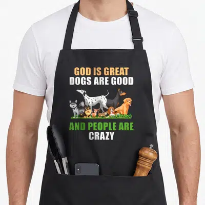 Funny god cooking with dog apron - Cartoon dogs on grass with text GOD IS GREAT DOGS ARE GOOD AND PEOPLE ARE CRAZY — shown on a black cotton canvas apron against a white background