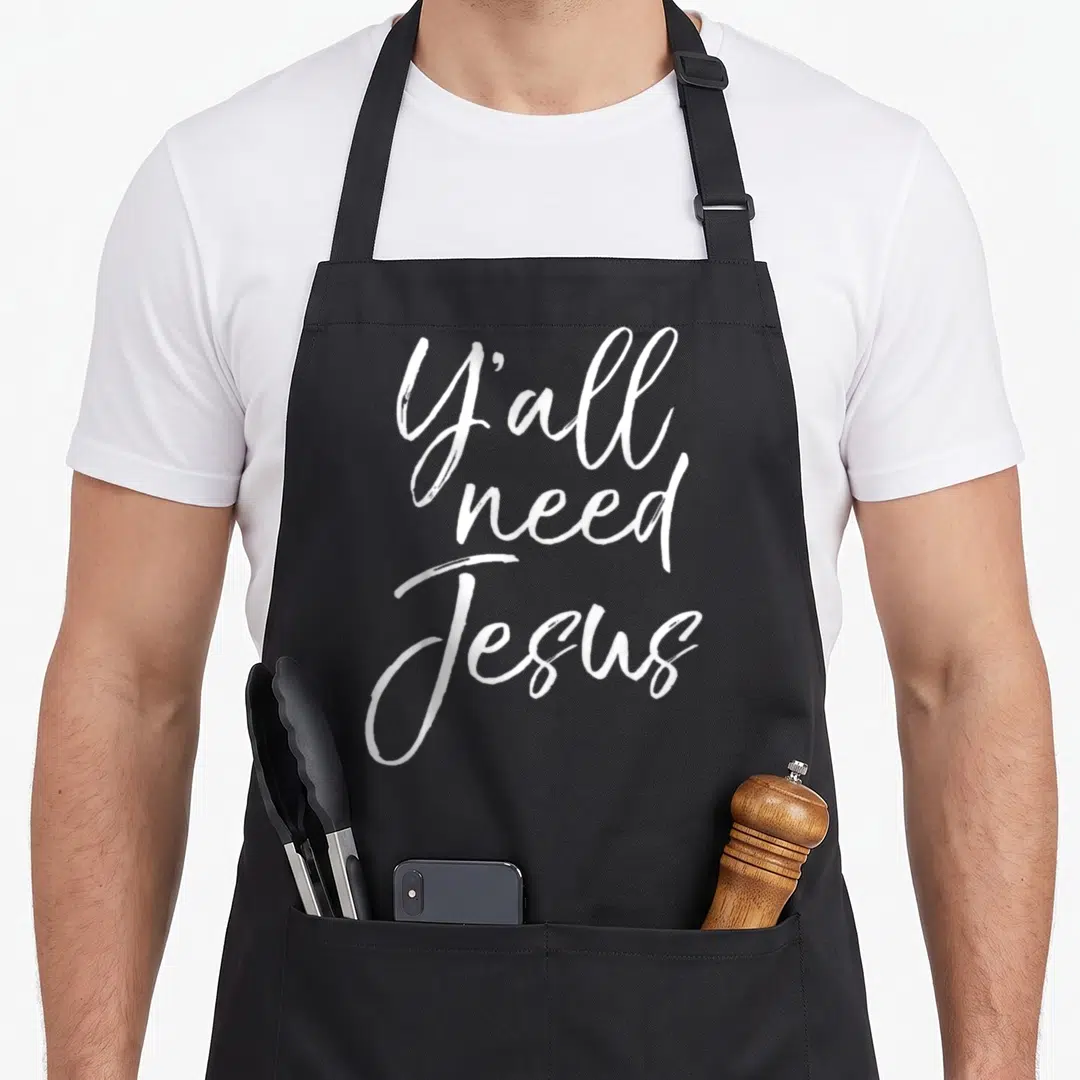 Jesus southern aprons - White stacked script text reading Y'all need Jesus — shown on a black cotton canvas apron against a white background