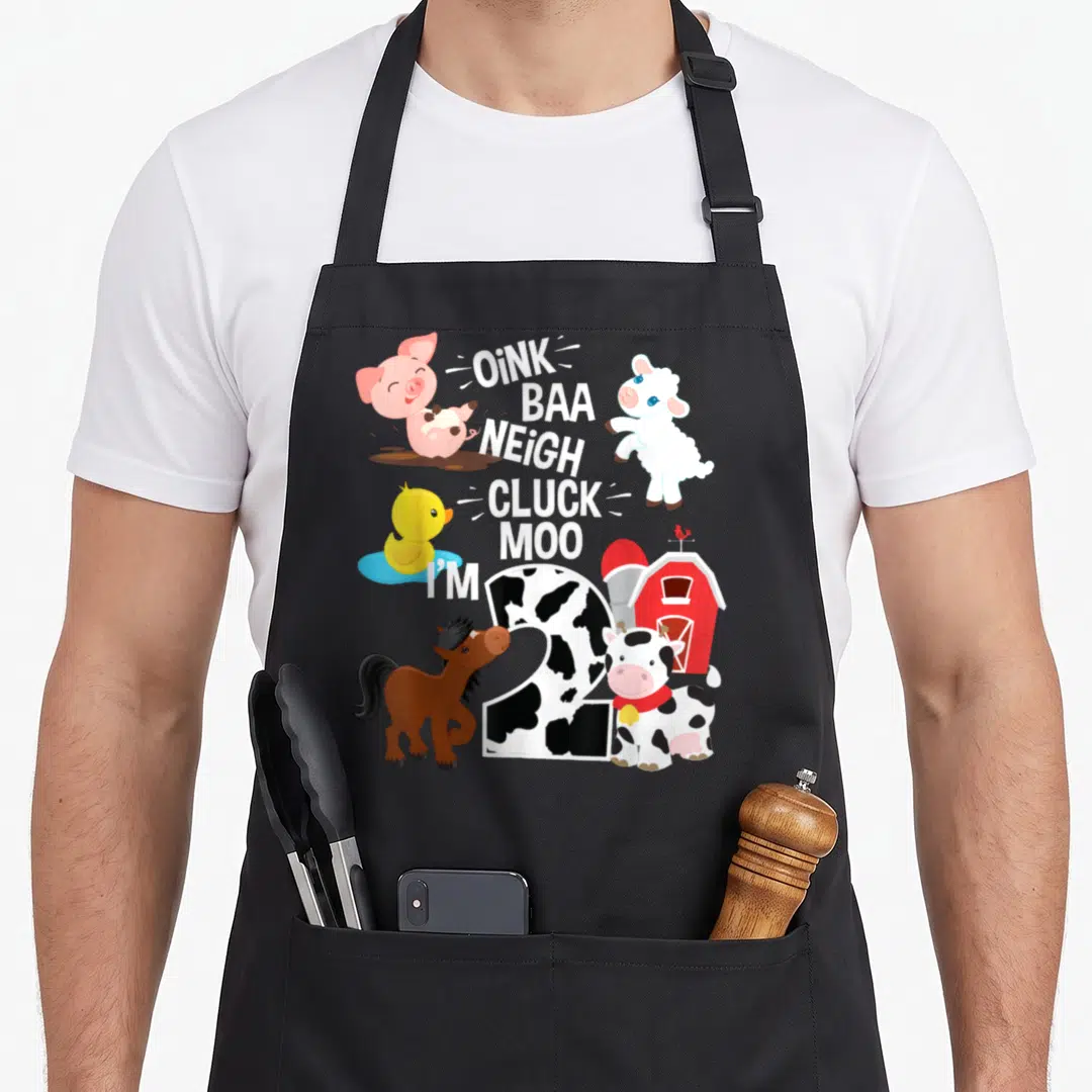 Baa farm apron - Cartoon number 2 with cow spots, farm animals, red barn, and stacked animal sound text — shown on a black cotton canvas apron against a white background