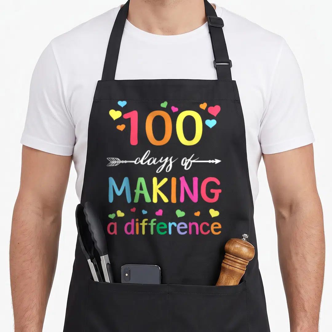 Difference making an apron - 100 days of making difference rainbow text with hearts and arrow graphic — shown on a black cotton canvas apron against a white background