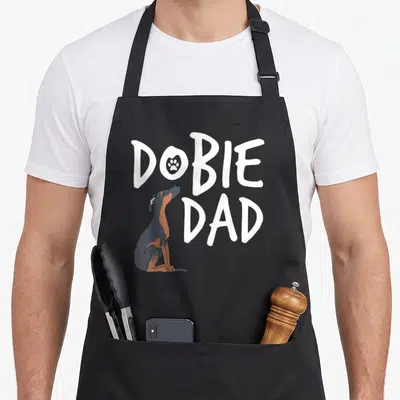 Pinscher puppy dog apron - White DOBIE DAD text with paw print in O and sitting grey tan dog — shown on a black cotton canvas apron against a white background