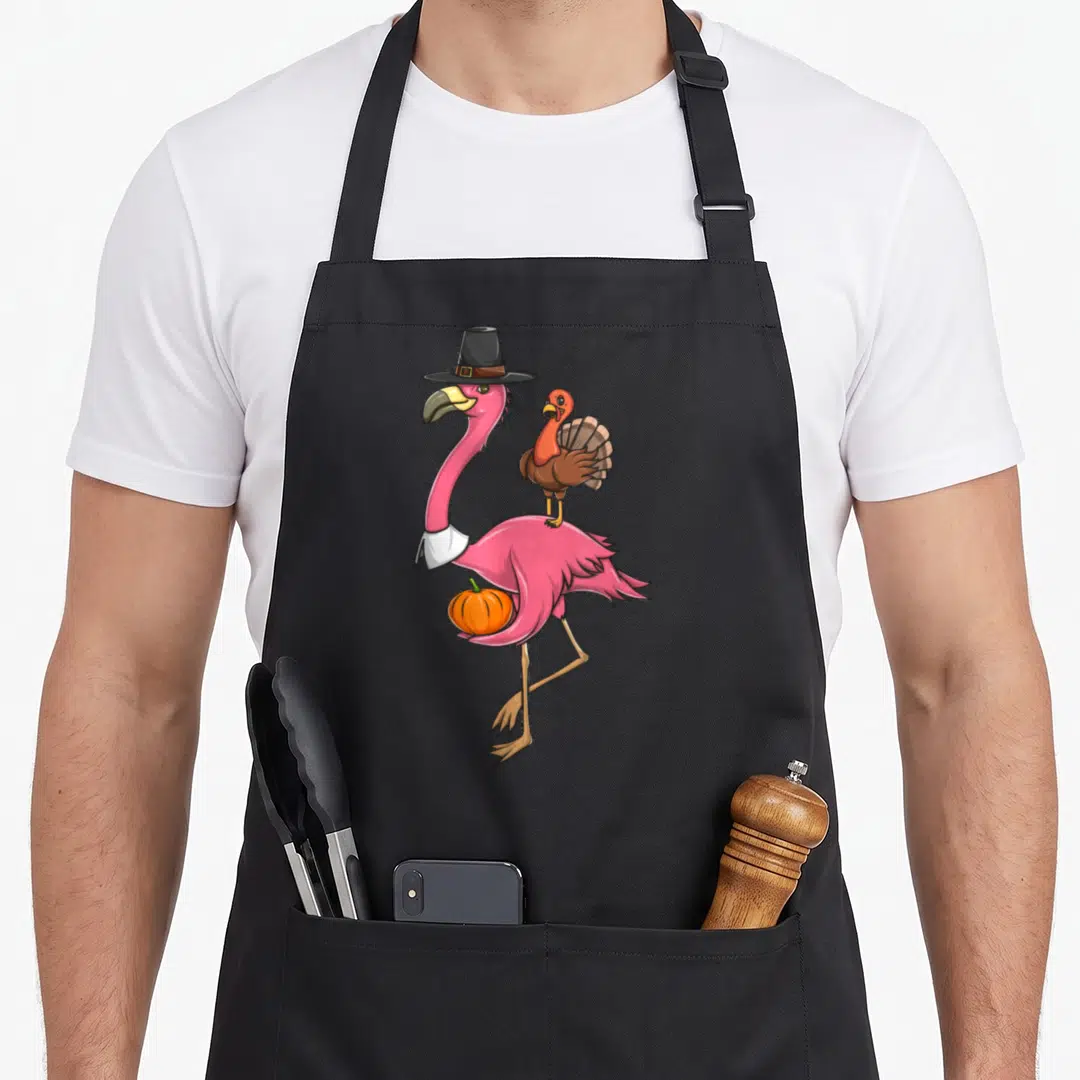 Flamingo pilgrim apron - Pink cartoon flamingo wearing a pilgrim hat holding a pumpkin with a turkey perched on its back — shown on a black cotton canvas apron against a white background