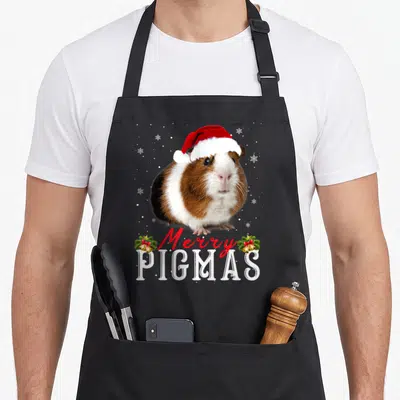 Pigmas merry christmas apron - Guinea pig in red santa hat with Merry PIGMAS text and two bell clusters — shown on a black cotton canvas apron against a white background