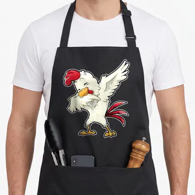 Dabbing chicken apron - Cartoon white rooster with red comb and yellow beak performing a dab dance pose — shown on a black cotton canvas apron against a white background