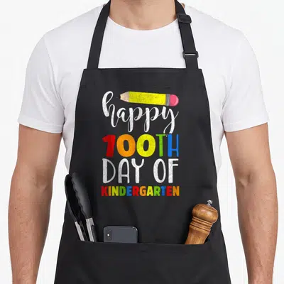 Funny 100th kindergarten teacher apron - Stacked happy 100th day of kindergarten text with yellow pencil — shown on a black cotton canvas apron against a white background