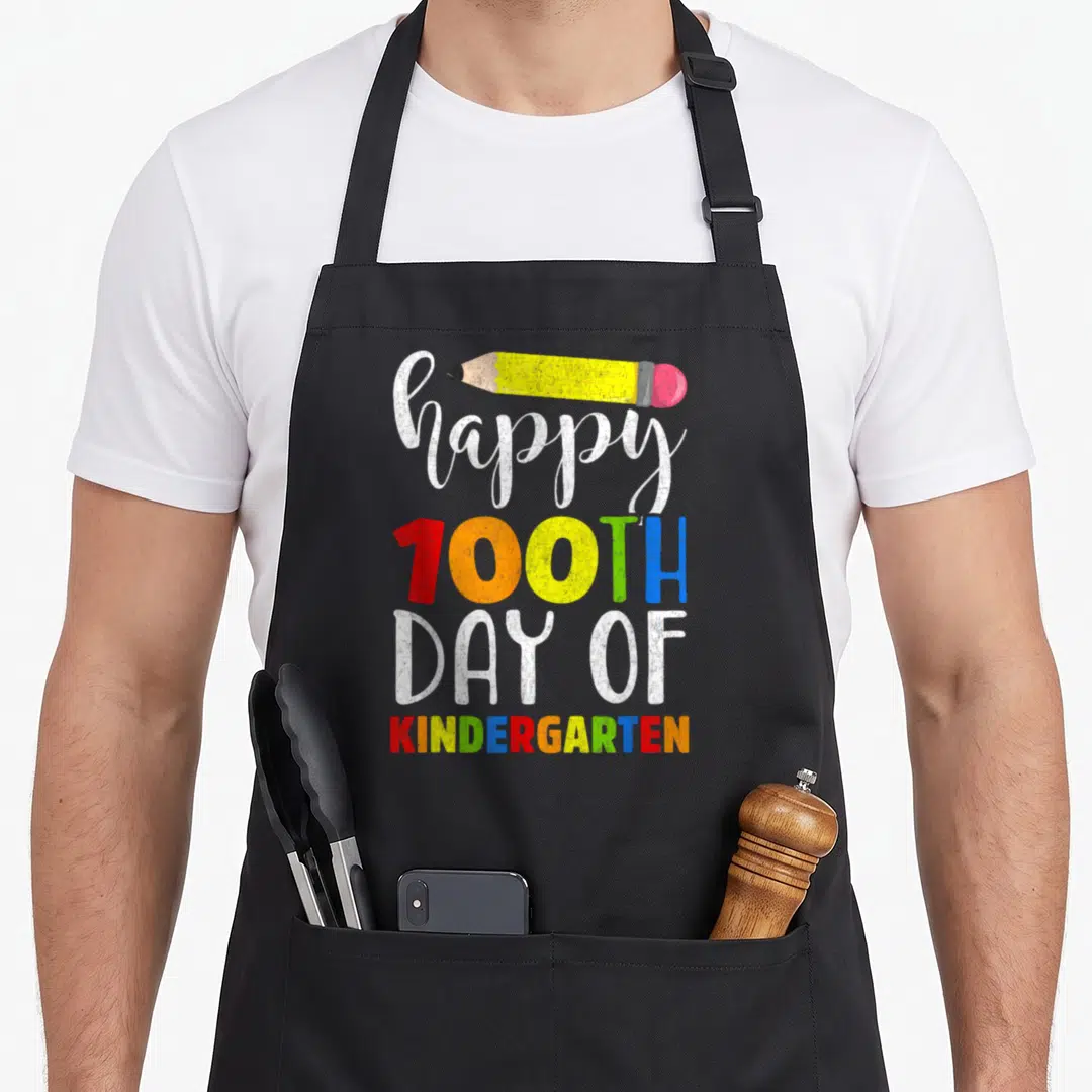Funny 100th kindergarten teacher apron - Stacked happy 100th day of kindergarten text with yellow pencil — shown on a black cotton canvas apron against a white background