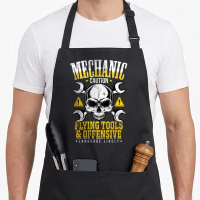 Caution auto mechanic apron - Skull crossed by two wrenches with yellow and white warning text — shown on a black cotton canvas apron against a white background