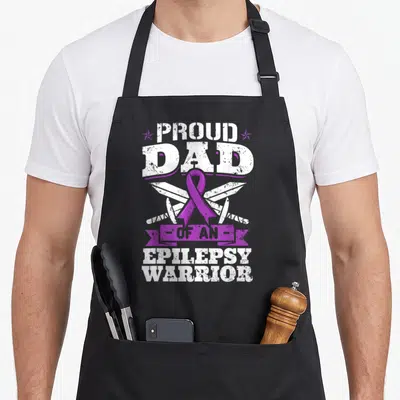 Proud epilepsy awareness apron - Purple ribbon over crossed white swords with text Proud Dad Of An Epilepsy Warrior — shown on a black cotton canvas apron against a white background