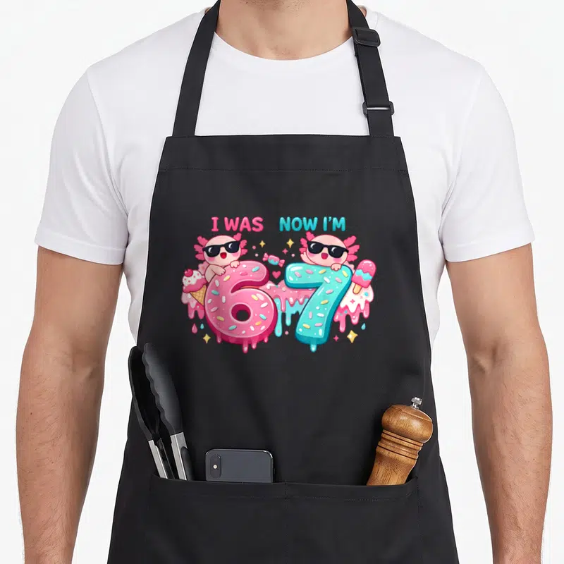 Axolotl meme apron - Two cartoon axolotls with sunglasses leaning on pink number 6 and blue number 7 surrounded — shown on a black cotton canvas apron against a white background