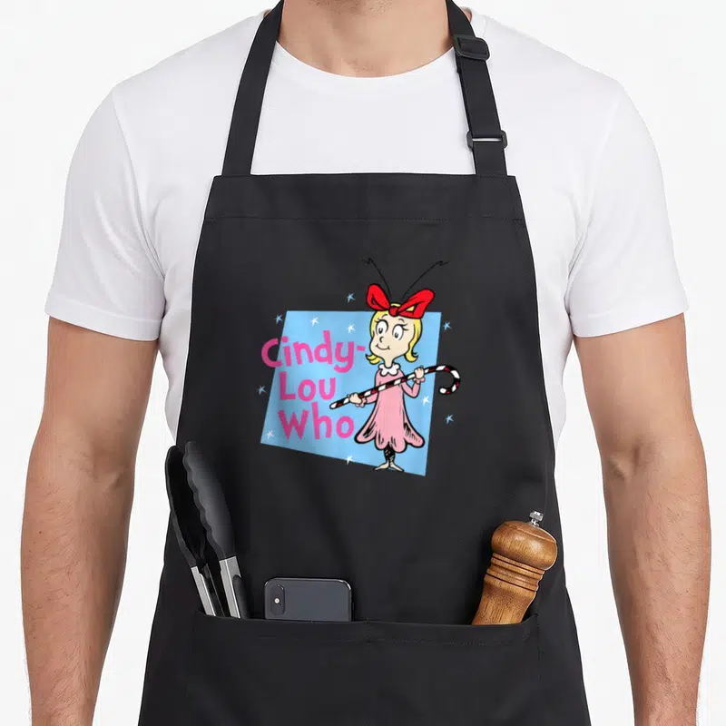 Funny holiday cindy lou who apron - Cindy-Lou Who character holding candy cane next to pink stacked text on blue — shown on a black cotton canvas apron against a white background