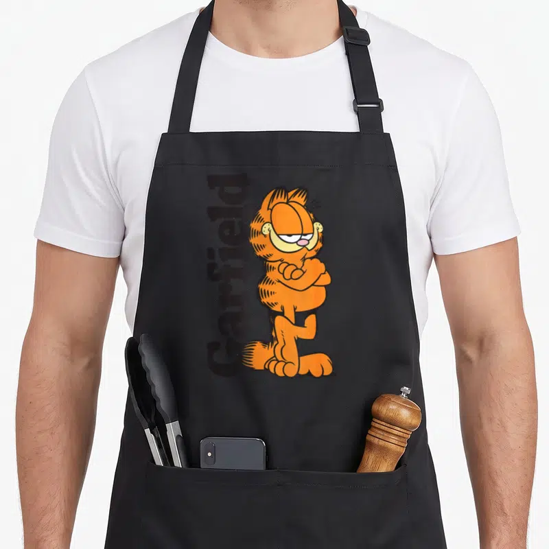 Leaning cat apron - Orange cartoon cat standing with arms crossed next to vertical black text reading Garfield — shown on a black cotton canvas apron against a white background