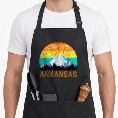 Sunset arkansas girl apron - Retro mountain sunset with orange yellow teal bands and ARKANSAS text below trees — shown on a black cotton canvas apron against a white background