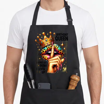 Older queen of baking apron - Gold crown on leopard lips with finger shush gesture and birthday queen text — shown on a black cotton canvas apron against a white background