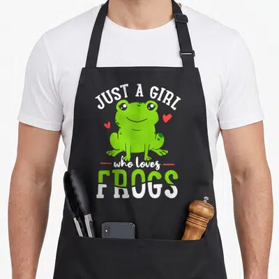 Cute animal frog aprons - Green cartoon frog with red hearts and text JUST A GIRL who loves FROGS — shown on a black cotton canvas apron against a white background