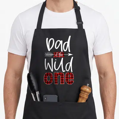 Wild the plaid apron - Dad of the Wild One text with red black plaid arrow and filled lettering — shown on a black cotton canvas apron against a white background