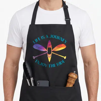 Journey kayak apron - Colorful kayak and crossed paddles with Life is a Journey Enjoyn the Ride text — shown on a black cotton canvas apron against a white background