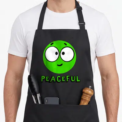 Version peaceful apron - Green cartoon smiley face with white eyes above black outlined text reading PEACEFUL — shown on a black cotton canvas apron against a white background