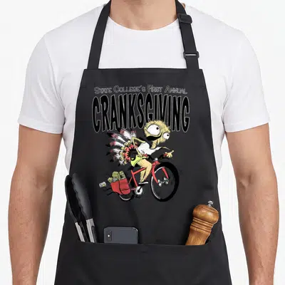 Cranksgiving state college apron - Stacked text above cartoon cyclist with turkey tail and red grocery bag — shown on a black cotton canvas apron against a white background
