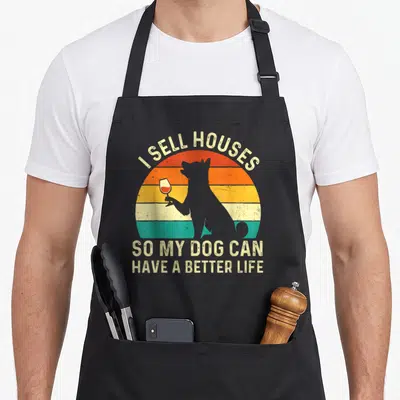 Estate dog apron - Grey dog silhouette holding wine glass against striped sunset with text I sell houses so my dog can — shown on a black cotton canvas apron against a white background